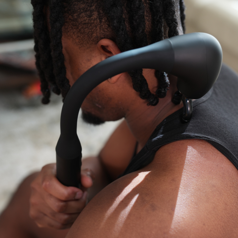 A person uses a black massage tool on their neck and shoulder. They are wearing a black tank top, sitting indoors with natural light, conveying relaxation. Backplus Hammer Massager - great too for athlete muscle recovery.