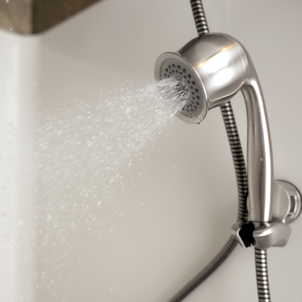 Close-up of a brushed nickel handheld showerhead in use, spraying water in a bright bathroom setting. The metal surface reflects light, emphasizing cleanliness.
