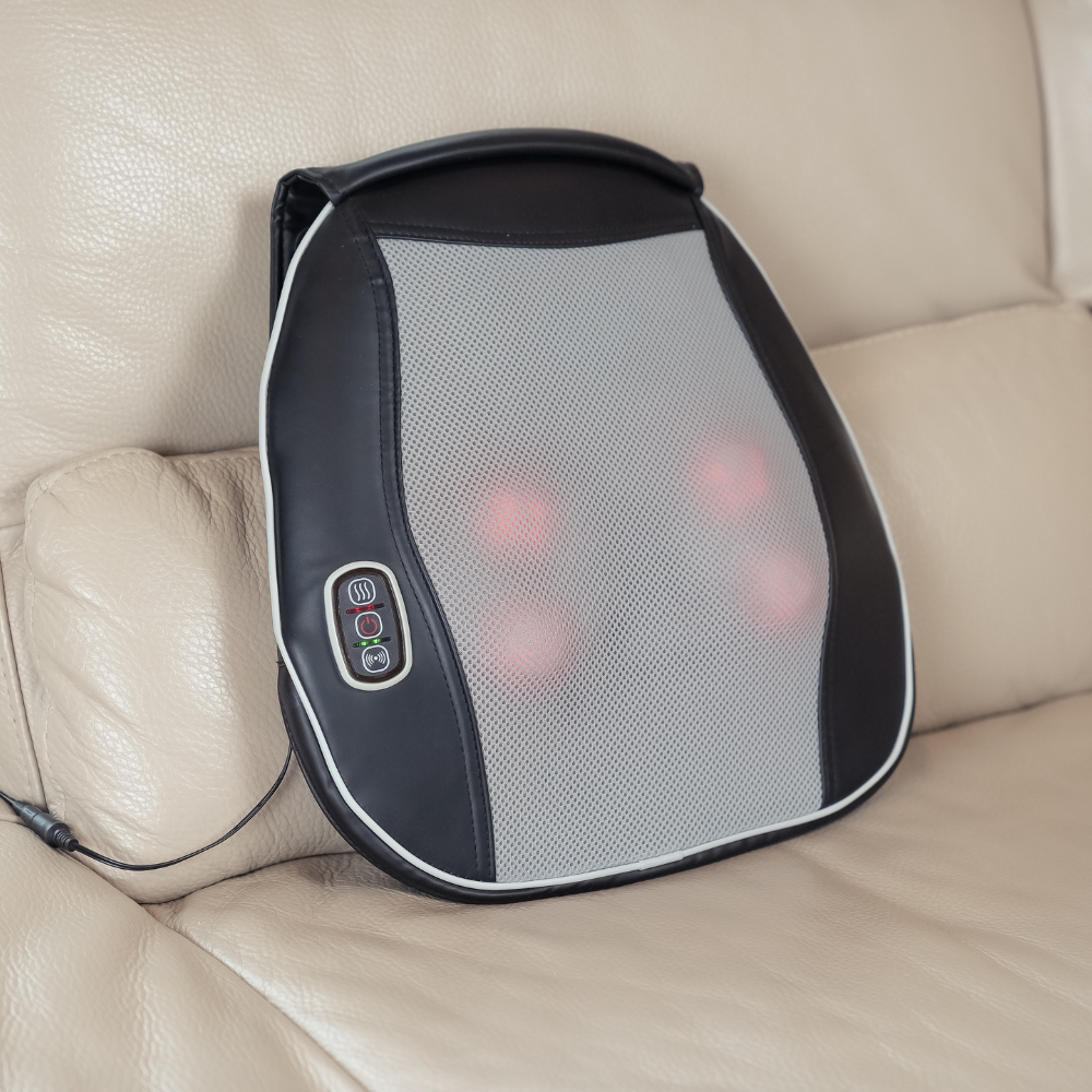 A black and mesh heated massager cushion rests on a cream leather sofa, featuring control buttons and glowing red massage nodes.