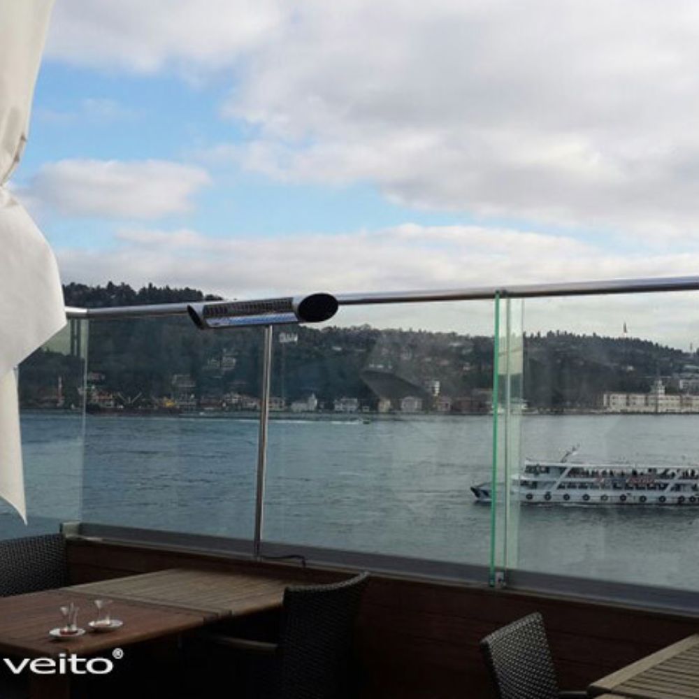 Veito heater on balcony with glass railings overlooking a body of water with a boat and cityscape in the background.