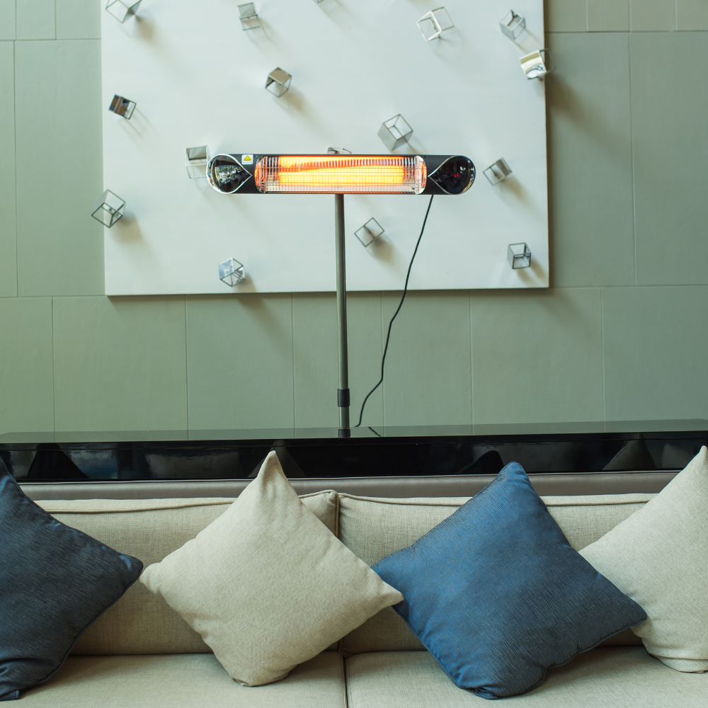 A modern infrared heater mounted on a stand, glowing orange, in front of a plush sofa with decorative pillows and an abstract wall art.