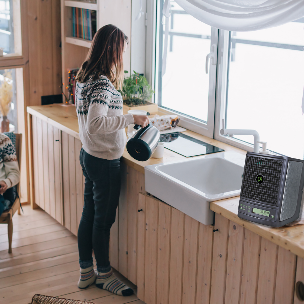 A woman in a cozy sweater pours water from a kettle into a bowl on a wooden kitchen counter, with snow outside the window and the iQ Pure Air™ 3000 Air Purifier on the counter.