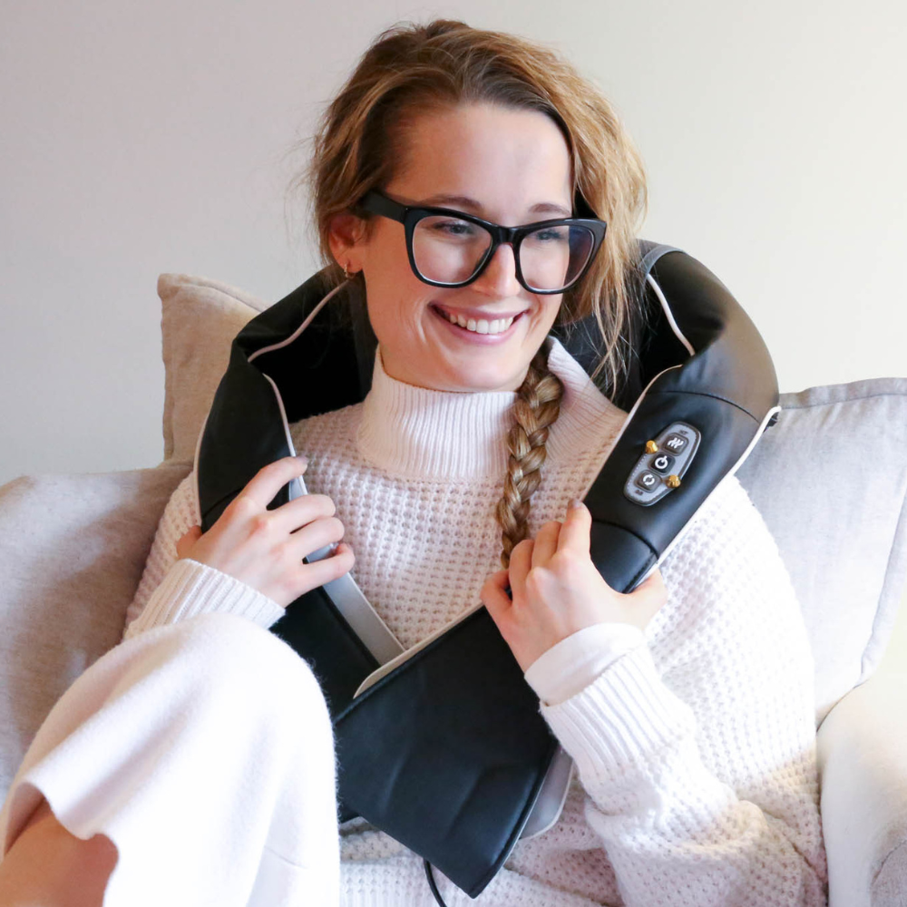 A person wearing a white knit sweater relaxes at home, holding a black neck massager with control buttons. Cozy atmosphere with soft furnishings.