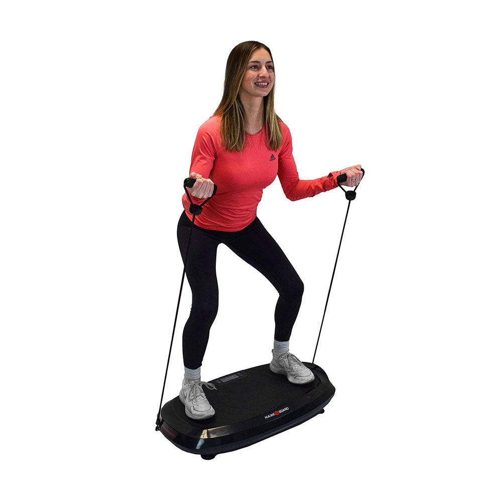 A woman exercises on a vibrating platform, holding resistance bands, wearing a red long-sleeve shirt and black leggings.