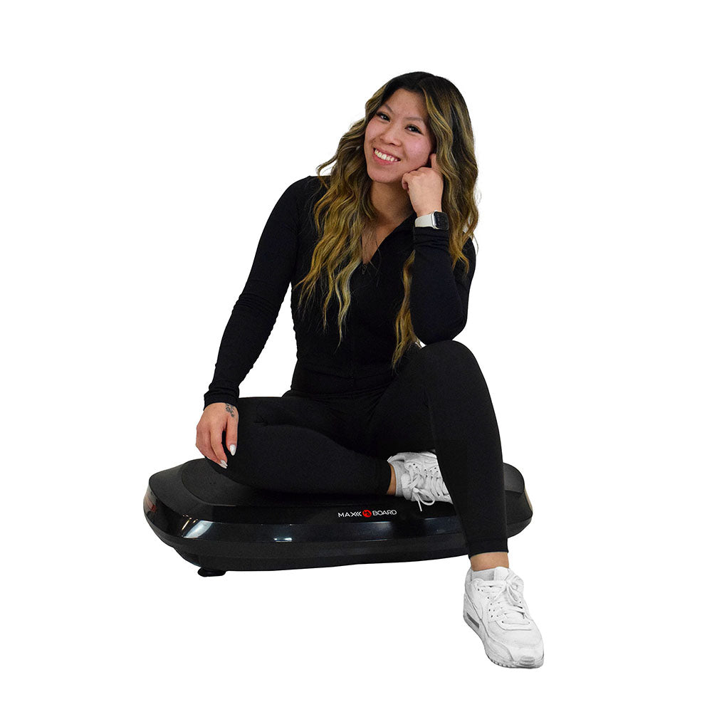A person in black athletic wear is sitting on a vibration plate, showcasing a fitness workout setting.