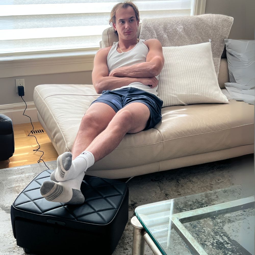 Man sitting on a beige couch with his legs crossed, wearing a white tank top and blue shorts, resting feet on a Backplus Ottoman Foot Massager