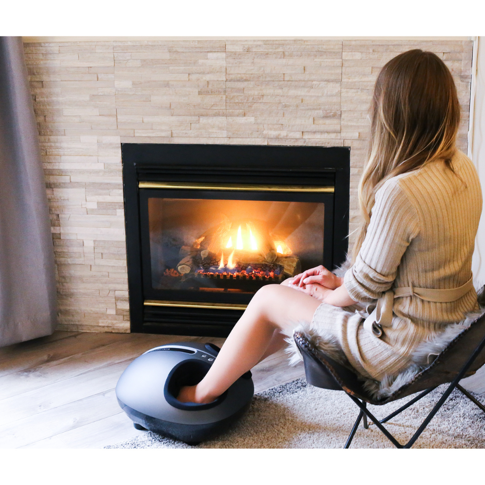 A woman relaxes in a cozy chair, enjoying a foot massager by a glowing gas fireplace, wrapped in a soft beige robe.