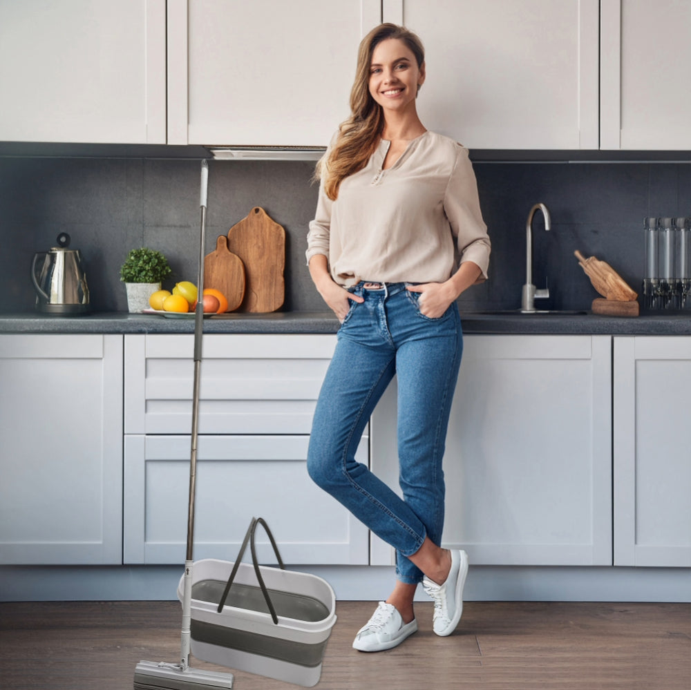 A woman smiles, leaning against a modern white kitchen counter. She wears casual jeans and a beige blouse. A mop and bucket are in the foreground.