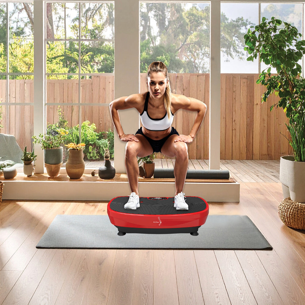 A woman in workout attire squats on a red vibration platform in a sunlit room with wooden floors and potted plants. The mood is focused and energetic.